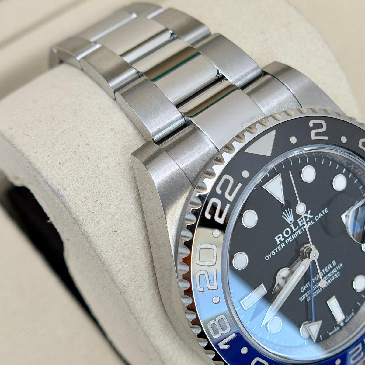 A close-up of a Rolex Oyster Perpetual Date watch with a stainless steel Oyster bracelet and a blue and black bezel, resting on a beige watch cushion.